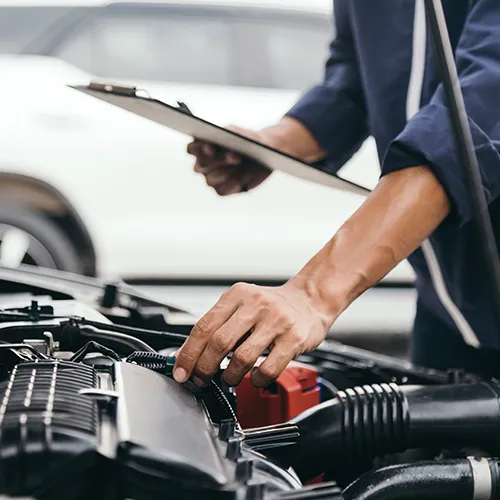 Mechanic holding a clipboard while performing a detailed under-hood vehicle inspection.